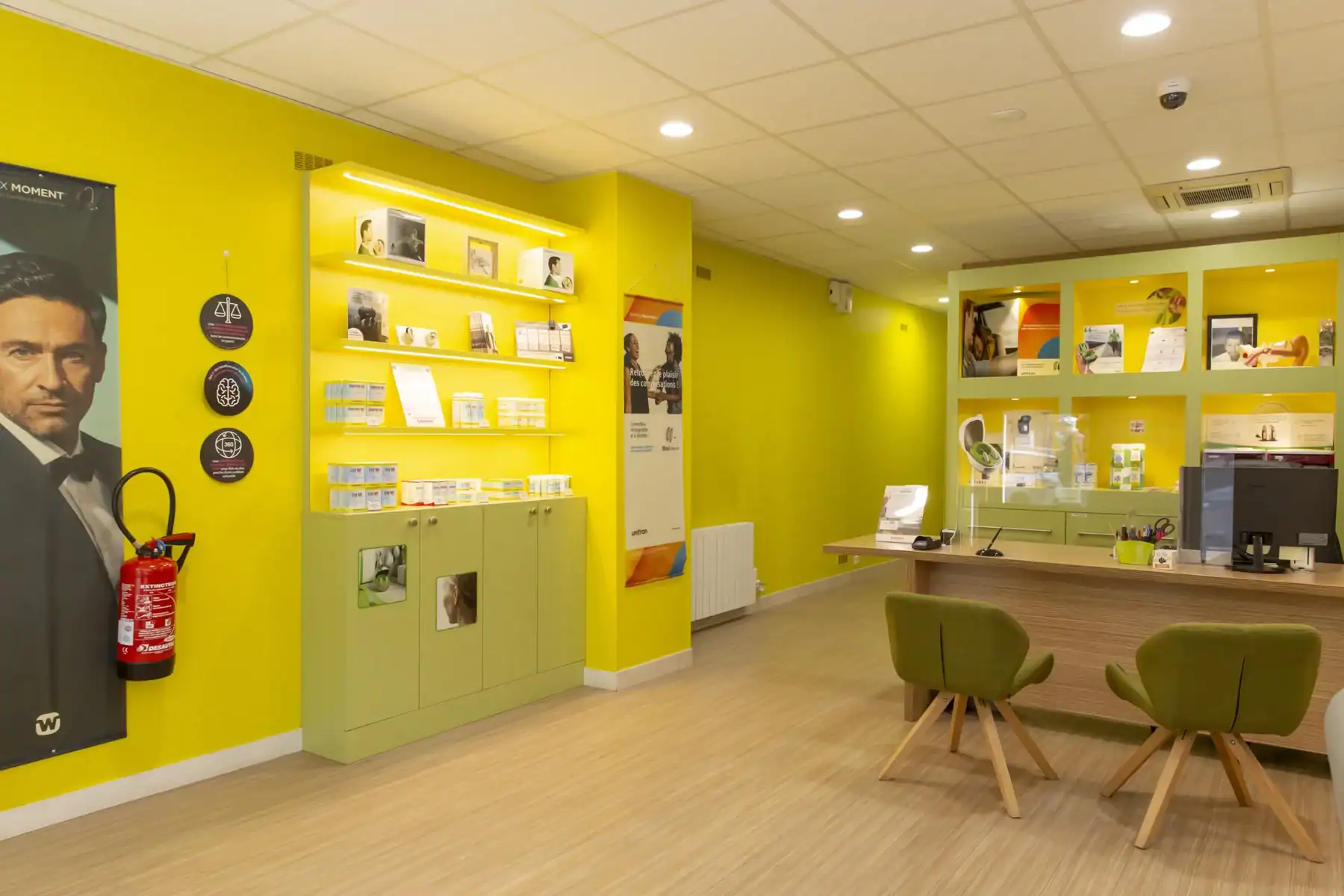 Espace d'accueil d'un centre d'audiologie avec des étagères de produits auditifs et un comptoir d'accueil lumineux en vert et jaune.
